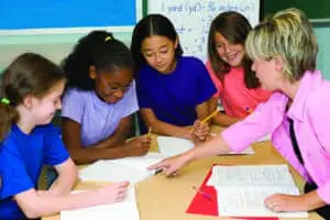 Students in a classroom. The teacher is pointing out what needs to be done in their notebooks.